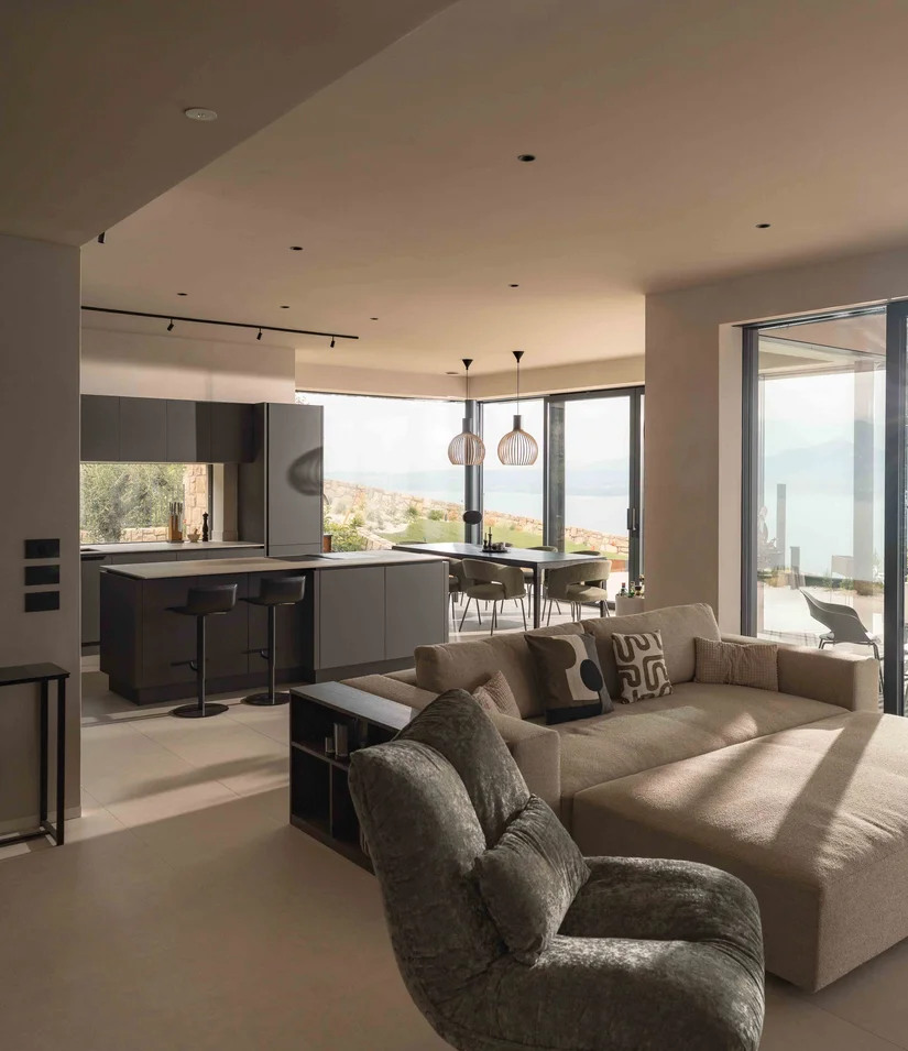 INTERIOS. Il tuo studio di interior design. Soggiorno moderno con cucina, divano e finestre panoramiche affacciate sul paesaggio e sul mare