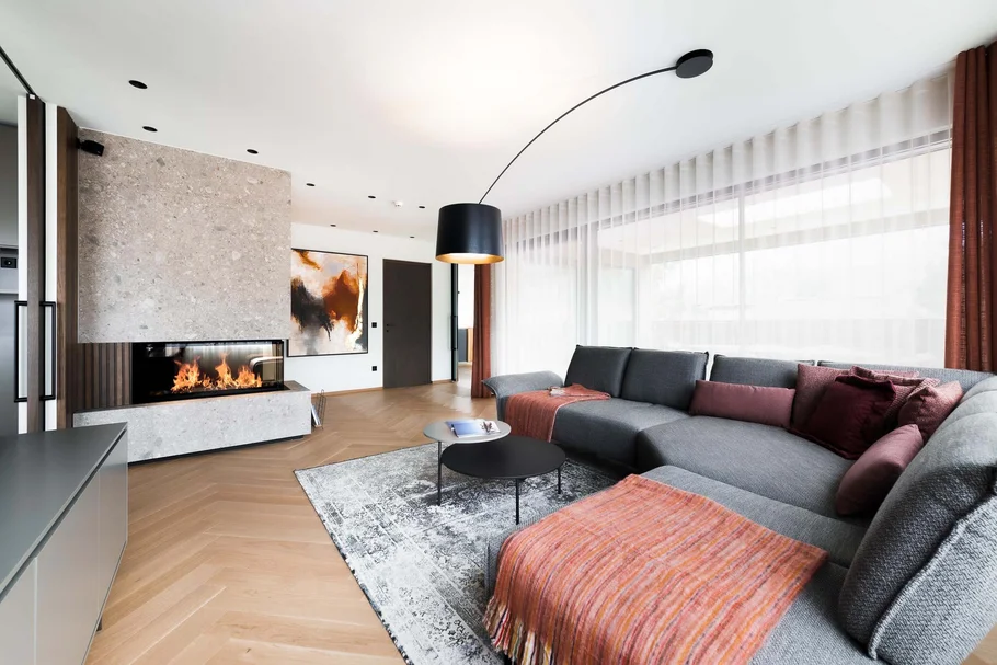 Moderner Wohnzimmer mit grauem Sofa, Kamin und großen Fenstern mit weißen Vorhängen