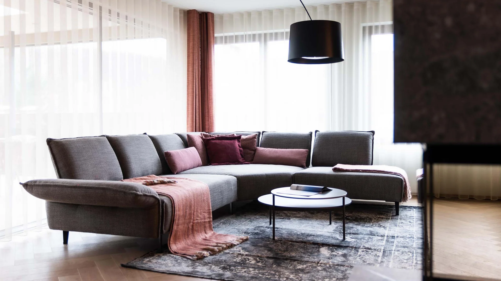 Helles Wohnzimmer mit grauem Ecksofa, rosa Kissen und Rundtisch