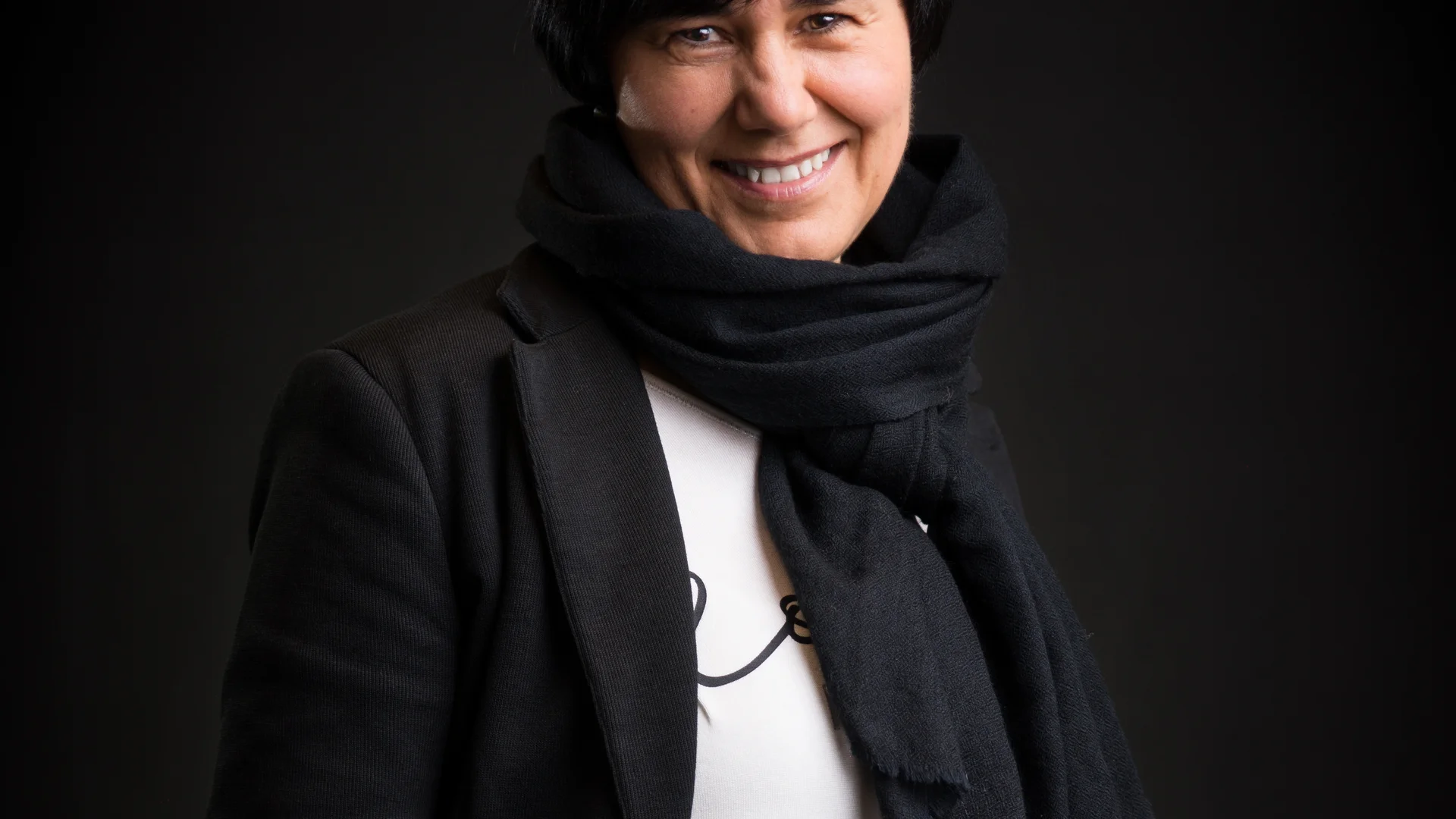 Lächelnde Person mit schwarzem Mantel und schwarzem Schal vor dunklem Hintergrund