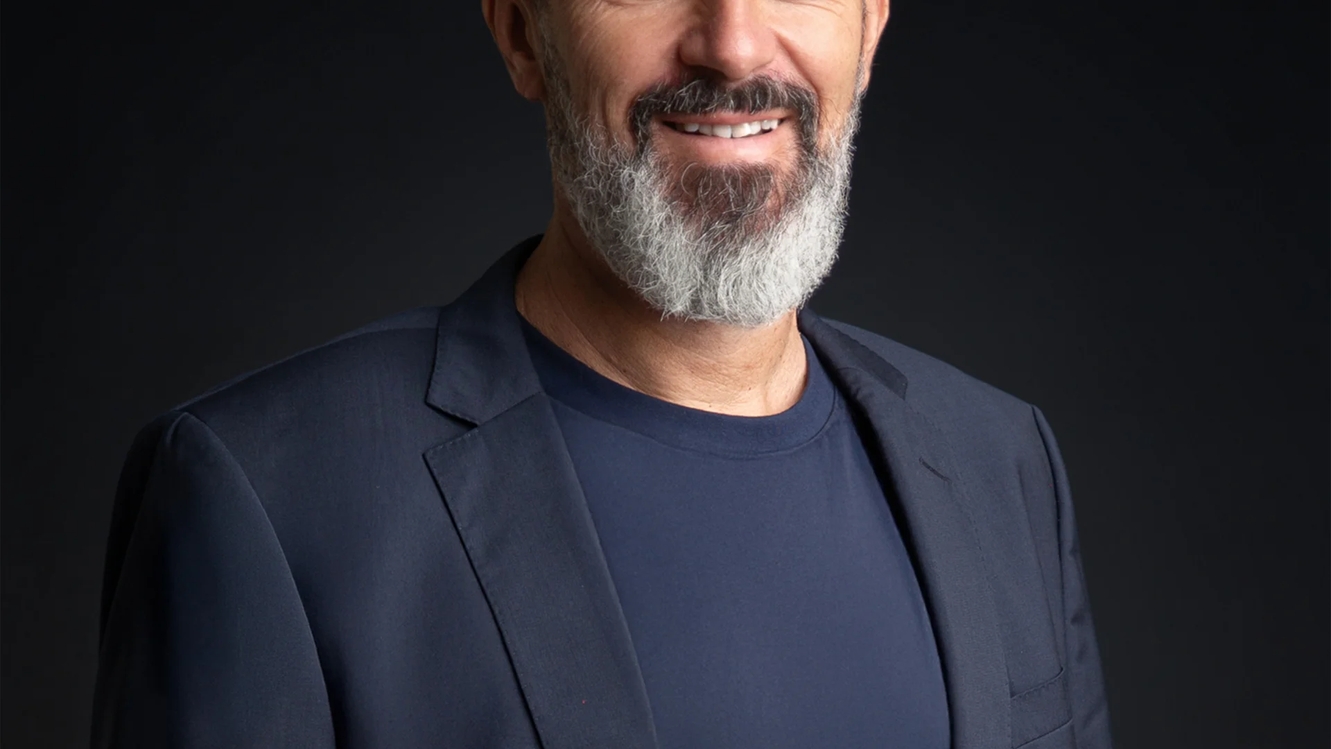 Lächelnder Mann mit grauem Bart in dunklem Anzug vor schwarzem Hintergrund