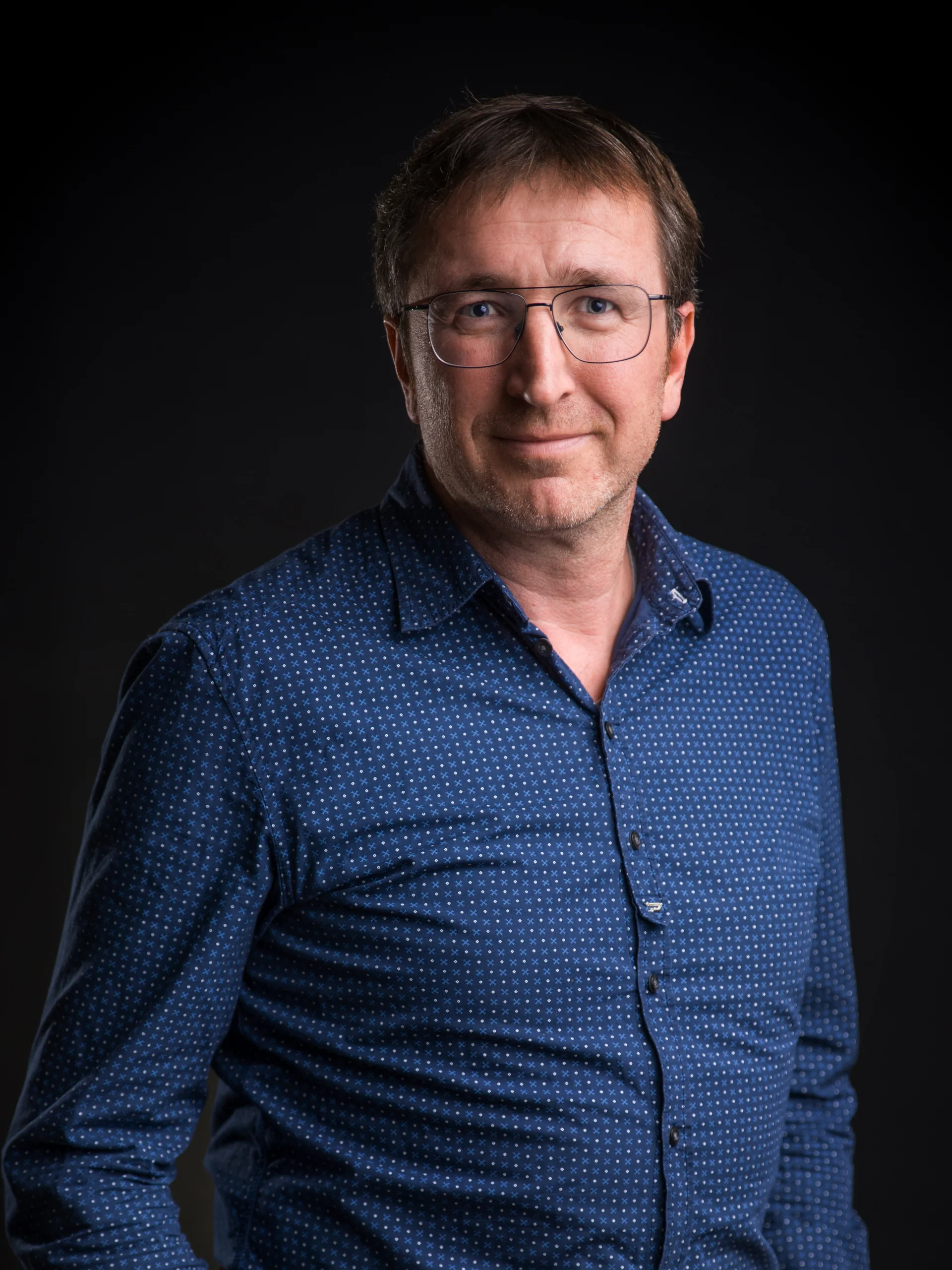 Mann mit Brille und blauem Hemd vor schwarzem Hintergrund