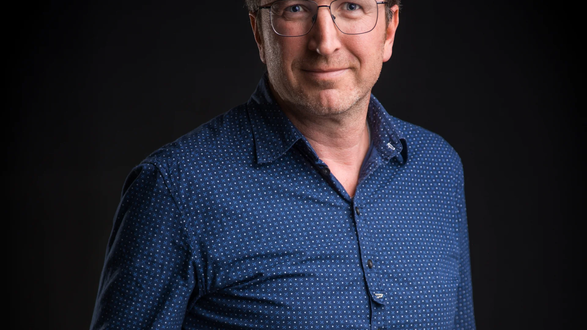 Mann mit Brille und blauem Hemd vor schwarzem Hintergrund