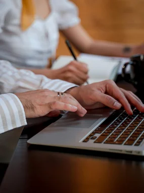 Persona che digita su laptop, altra persona scrive su un taccuino sullo sfondo