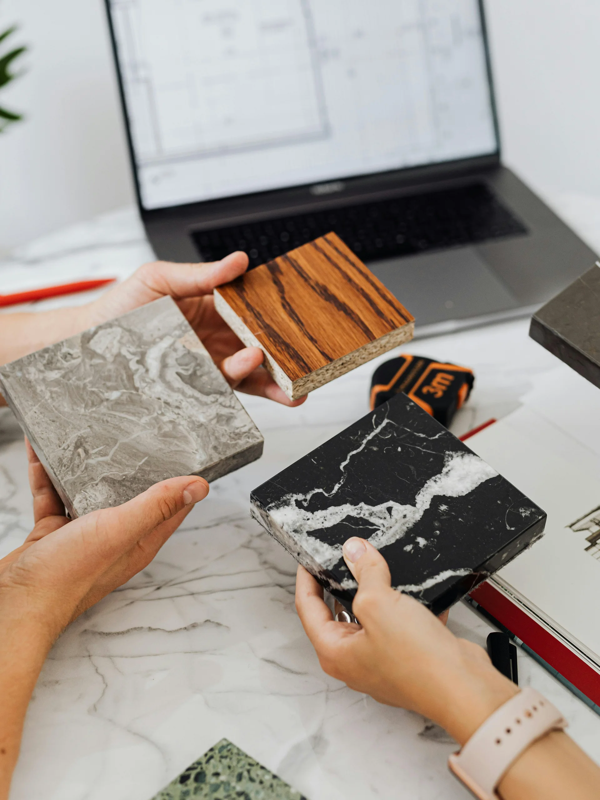 Personen vergleichen verschiedene Holz- und Steinproben vor einem Laptop mit Architekturplänen