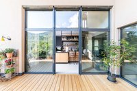 Blick durch große Glasfenster auf eine moderne Küche mit Holzterrasse und Pflanzen