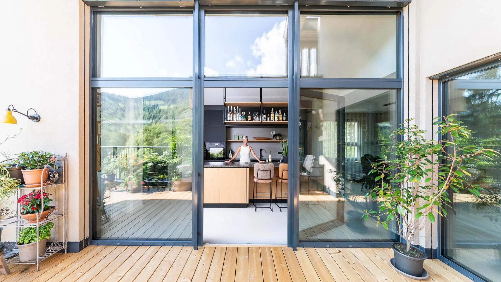 Blick durch große Glasfenster auf eine moderne Küche mit Holzterrasse und Pflanzen