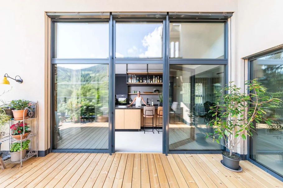 Blick durch große Glasfenster auf eine moderne Küche mit Holzterrasse und Pflanzen